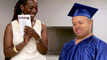 Special education student in graduation outfit, smiling next to woman clapping 