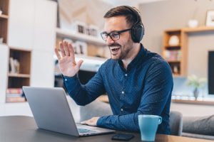 A behavior analyst wearing headphones waves and smiles during a telehealth ABA therapy session on a laptop, home office setting