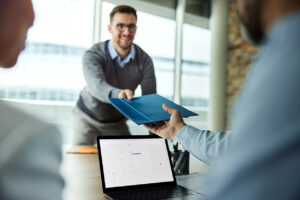 A smiling man in glasses passes a portfolio folder across a desk to a colleague, with a laptop open in the foreground