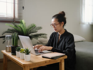 A woman in glasses types on a laptop at a wooden desk with small potted plants, researching ABA degree programs from home
