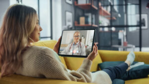 Person relaxing on a couch holding a tablet during a telehealth video call with a healthcare professional at home