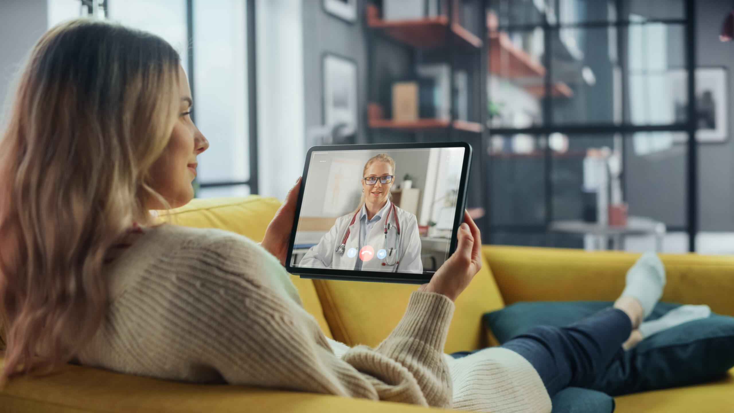 Woman on a couch holding a tablet on a video call with a health professional