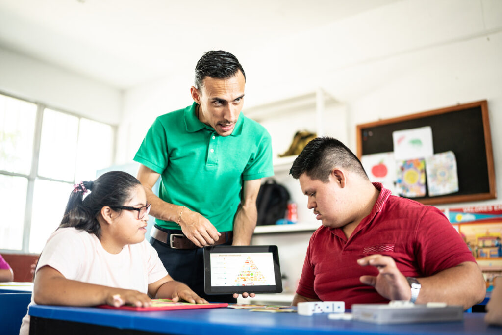 Male teacher holding a tablet and explaining a lesson about the food pyramid to two students, both of whom have Down Syndrome