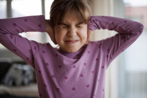 A young girl in a purple shirt grimaces and covers both ears tightly with her hands while standing near a window indoors.