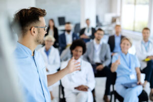 A healthcare professional in scrubs gestures while presenting to a seated audience of medical staff in a bright conference room.